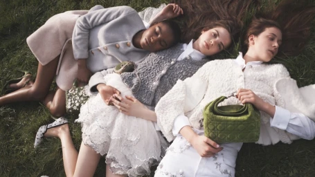 Four models lying on green grass outdoors holding small green handbags during Dior Fall-Winter 2026 campaign.