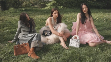 Three female models sitting on a lush green lawn, wearing designer dresses and coats while posing with luxury handbags.