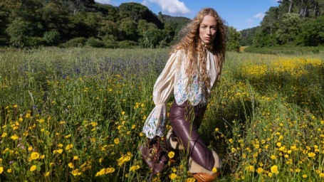 Model with long blonde curly hair standing in a vibrant yellow wildflower field wearing an embroidered ivory blouse and dark leather trousers