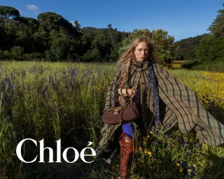 Model standing in a grassy meadow wearing a plaid cape and holding a brown leather handbag, with a forest background under a clear sky.