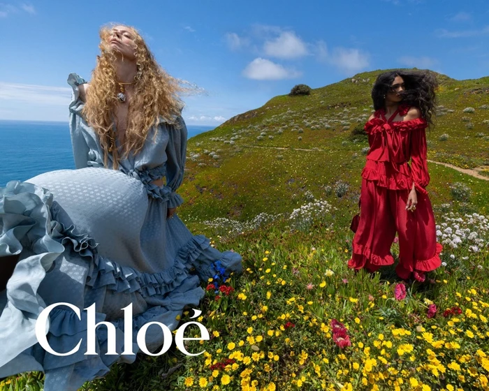 Two models in ruffed dresses standing in a wildflower field by the sea for Chloé Fall-Winter 2026 campaign.