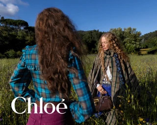 Two women with long curly hair standing in a green meadow wearing plaid apparel for the Chloé Fall-Winter 2026 campaign.