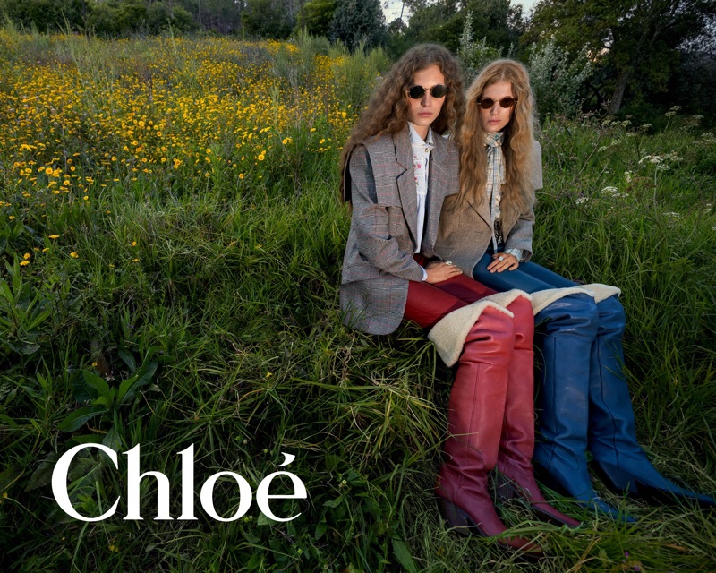 Two female models in blazers and over-the-knee boots seated in a grassy meadow filled with yellow wildflowers.