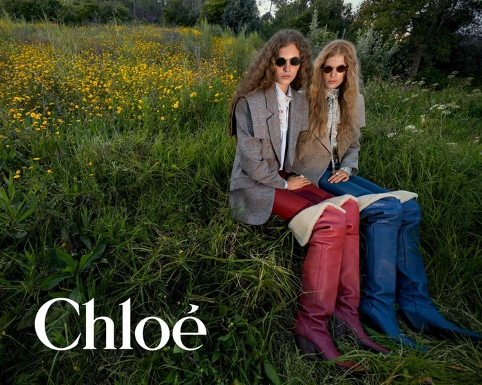 Two female models in blazers and over-the-knee boots seated in a grassy meadow filled with yellow wildflowers.
