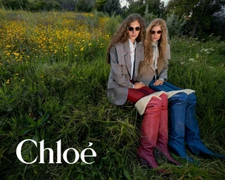 Two female models in blazers and over-the-knee boots seated in a grassy meadow filled with yellow wildflowers.