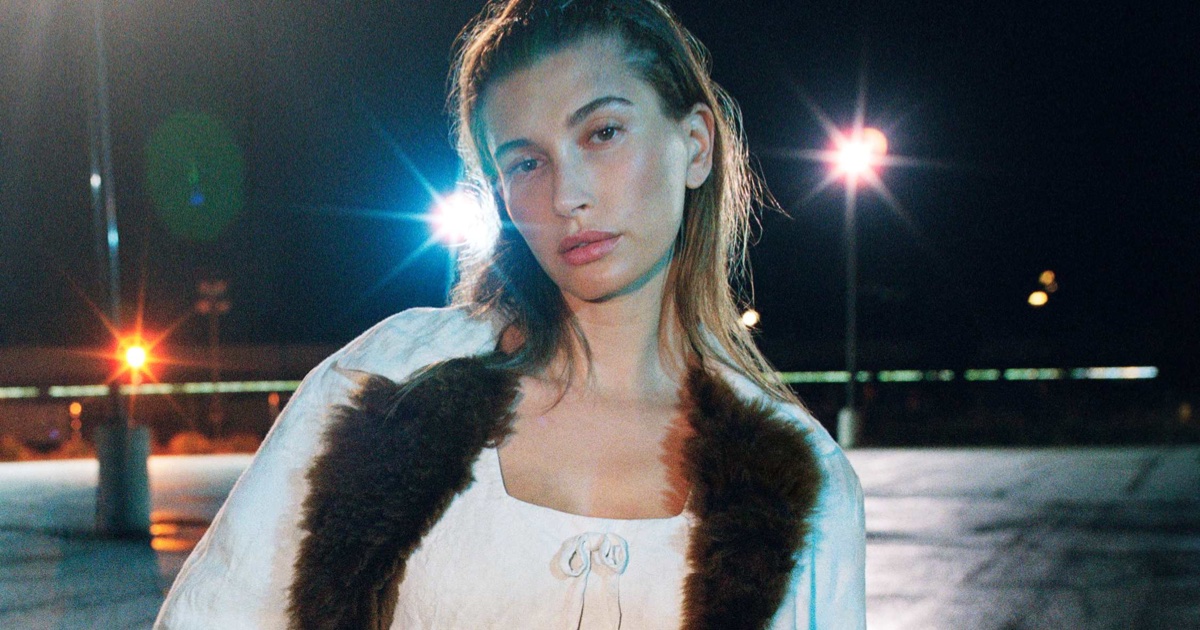 Model Hailey Bieber posing outdoors at night under streetlights wearing a light-coloured top with dark fur trim.