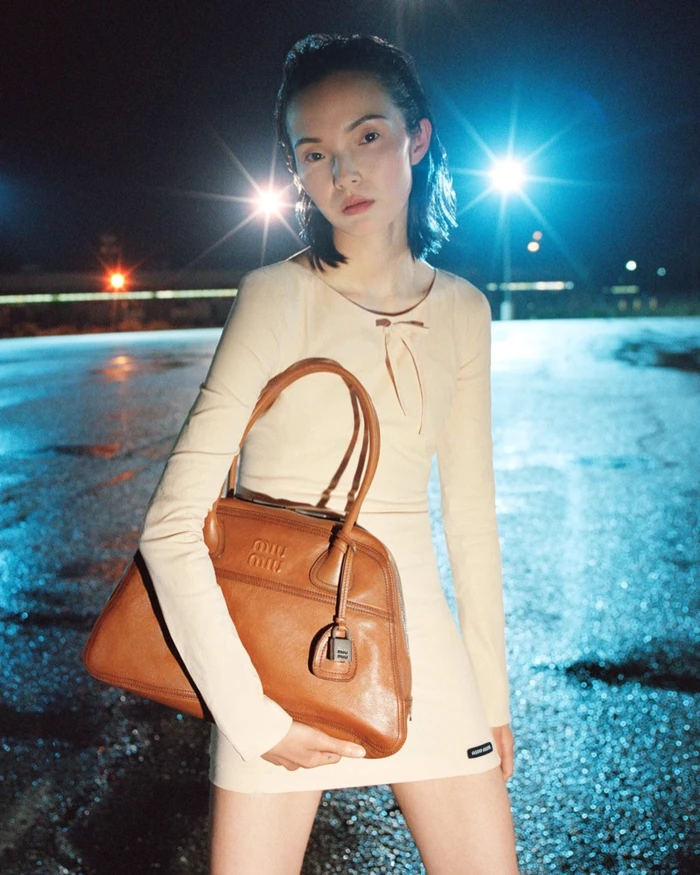 Xiao Wen Ju holding a brown leather handbag outdoors at night under bright streetlights for Miu Miu Fall-Winter 2026 Campaign