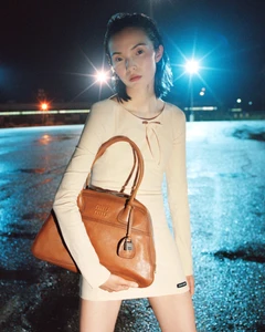 Xiao Wen Ju holding a brown leather handbag outdoors at night under bright streetlights for Miu Miu Fall-Winter 2026 Campaign