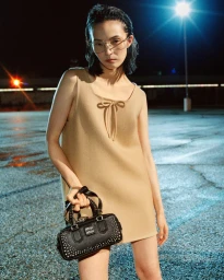 Xiao Wen Ju in a beige dress holding a black studded handbag outdoors at night under bright stadium lights for the Miu Miu Fall-Winter campaign