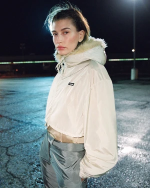 Hailey Bieber wearing a cream hooded puffer jacket and grey trousers standing outdoors at night for Miu Miu.