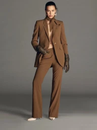 Model Irina Shayk wearing a brown pantsuit, matching neck scarf, and dark leather gloves against a plain grey background