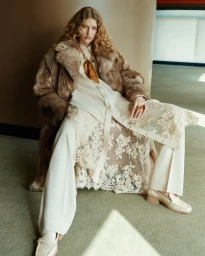 Model with long curly hair wearing a fluffy brown fur coat, cream lace tunic, white trousers, and loafers, seated indoors.