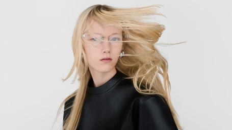 Model with long blonde hair blowing in wind wearing frameless glasses and black top against white background