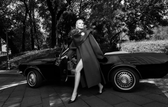 Nadja Auermann in a long structured coat stepping out of a vintage convertible on a paved street.