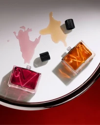 Two open glass Valentino perfume bottles with magenta and amber liquid beside matching liquid spills on a white glossy surface.
