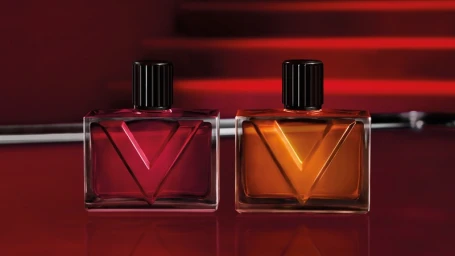 Two rectangular glass perfume bottles with ribbed black caps on a dark red reflective surface with a blurred background