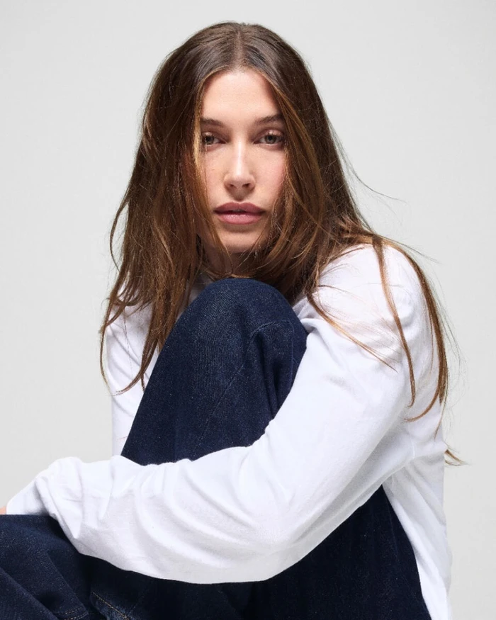 Hailey Bieber wearing a white long-sleeve top and dark denim jeans against a plain white background for Gap by Mario Sorrenti.