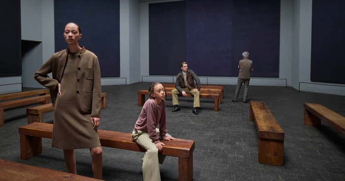 Model in brown jacket and matching dress standing on wooden benches inside a gallery space for Loro Piana Fall-Winter 2026 Campaign