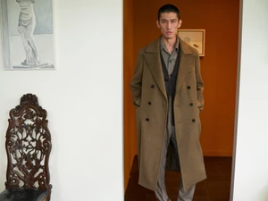 Man wearing a brown double-breasted coat over a grey shirt and dark vest standing in an interior space