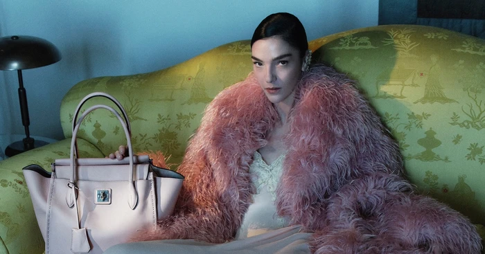Model Mariacarla Boscono in a pink feather coat seated on a patterned green sofa holding a pink leather handbag