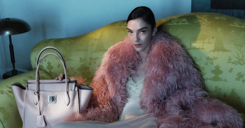 Model Mariacarla Boscono in a pink feather coat seated on a patterned green sofa holding a pink leather handbag