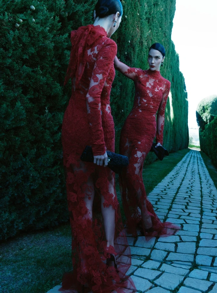 Model Mariacarla Boscono in a red lace gown holding a black clutch walking along a stone path beside tall green hedges.
