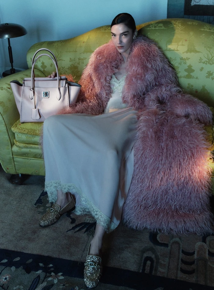 Model Mariacarla Boscono seated on a green sofa wearing a pink feather coat and holding a pink leather handbag