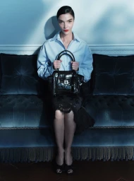 Mariacarla Boscono seated on a dark tufted sofa holding a black leather handbag for Ermanno Scervino Fall-Winter 2026 campaign