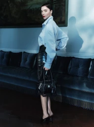 Model Mariacarla Boscono in a light blue shirt and dark textured skirt holding a black handbag in an interior setting.