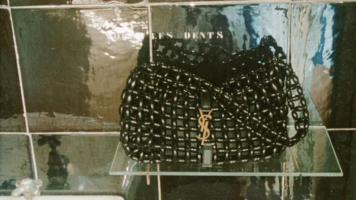 Black woven leather Yves Saint Laurent shoulder bag with a gold YSL logo resting on a glass shelf against dark green tiled walls.