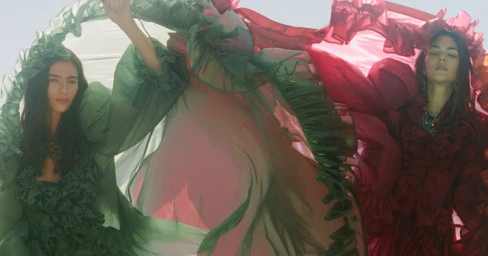 Two models in flowing green and red ruffled sheer garments holding large draped fabrics outdoors against a bright sky.