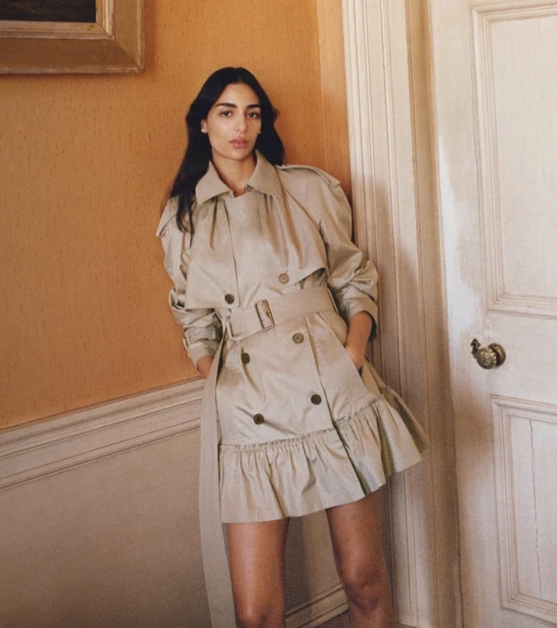 Model wearing a beige belted trench coat leaning against a warm yellow interior wall next to a white door.