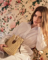 Model in white shirt holding a straw woven bucket bag against floral wallpaper for Dior