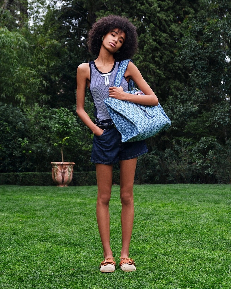 Woman with afro hair wearing a striped tank top and denim shorts holding a blue patterned tote bag in a garden