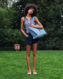 Woman with afro hair wearing a striped tank top and denim shorts holding a blue patterned tote bag in a garden