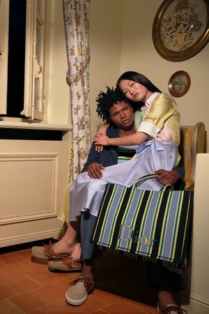 Two models in a warm interior setting holding a striped Dior tote bag for the Dioriviera Summer 2026 campaign.
