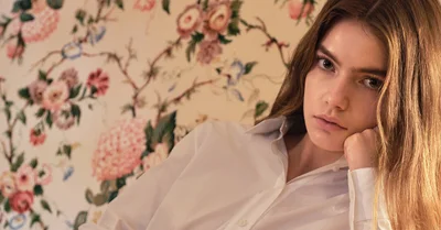 Woman with long brown hair wearing a white collared shirt resting her head on her hand against a floral patterned wallpaper background