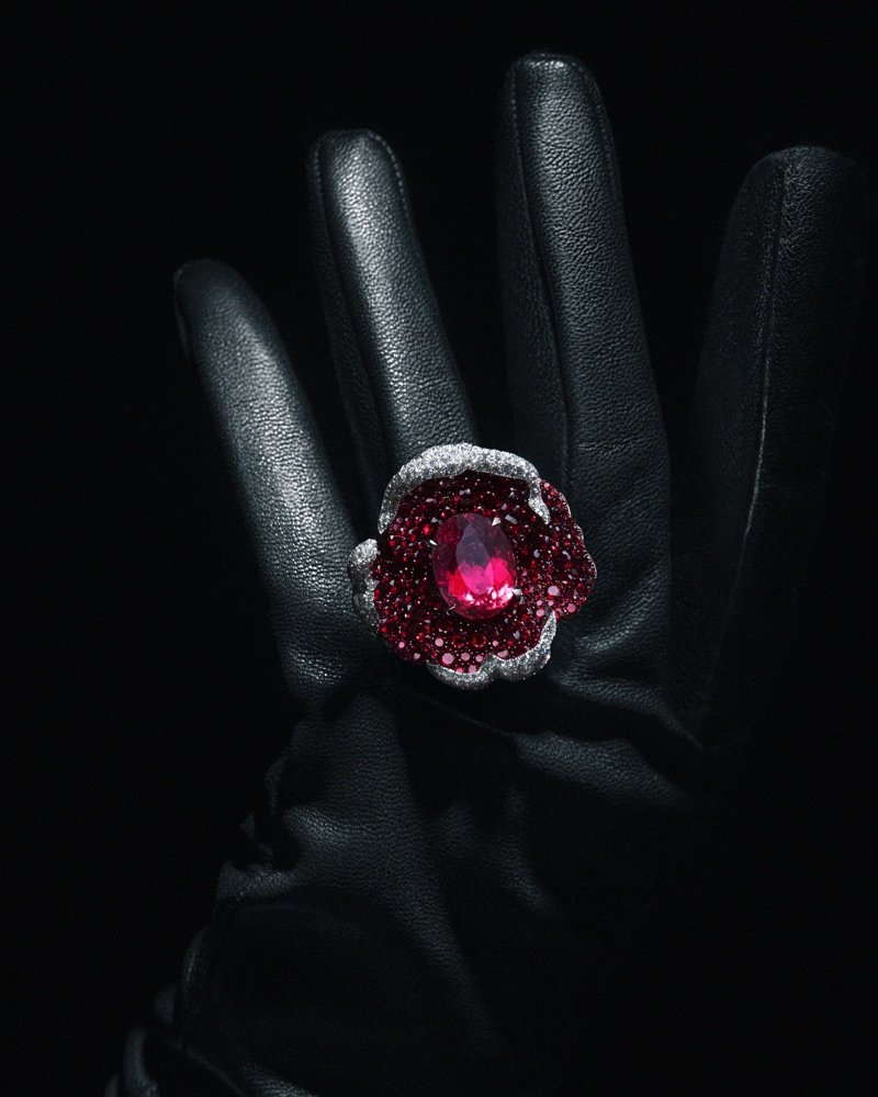 Close-up of a floral statement ring with a ruby center and diamond trim worn on a hand in a black leather glove against a dark background.