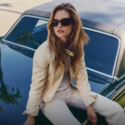 Model wearing dark sunglasses and a beige jacket seated on the hood of a dark car against a bright background.