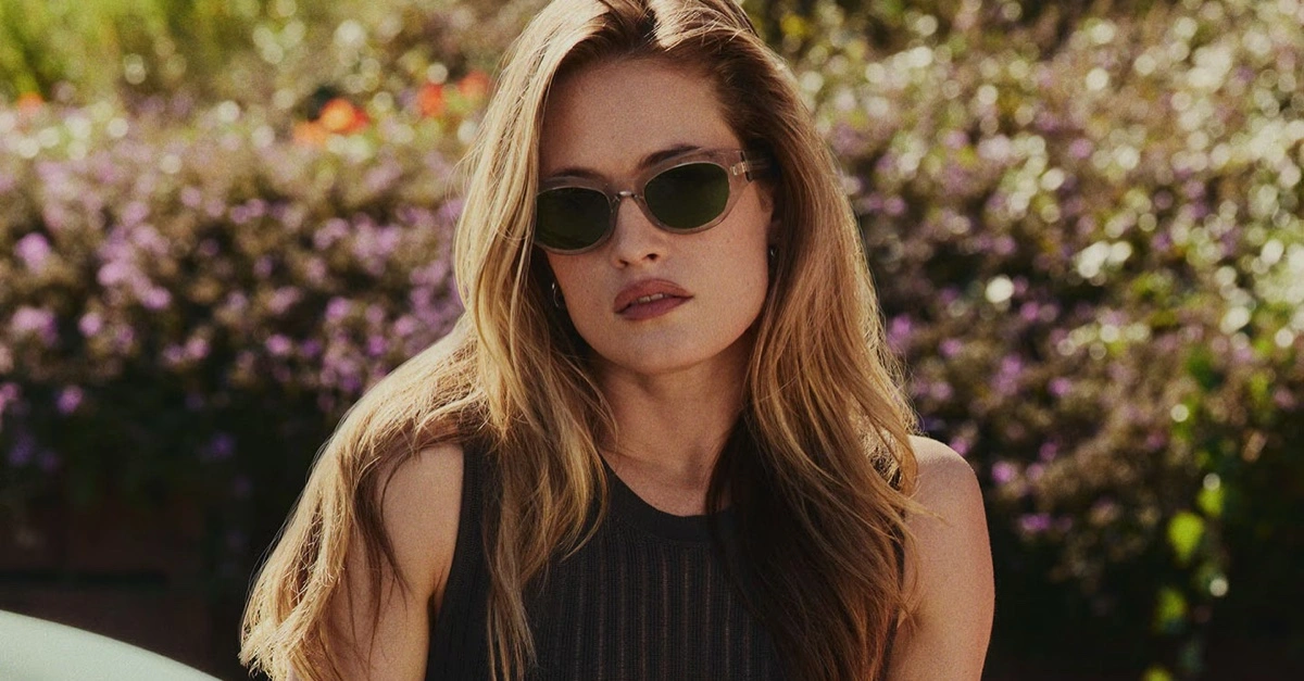 Model Hailey Carlson wearing sunglasses outdoors against a blooming floral background for Oliver Peoples.