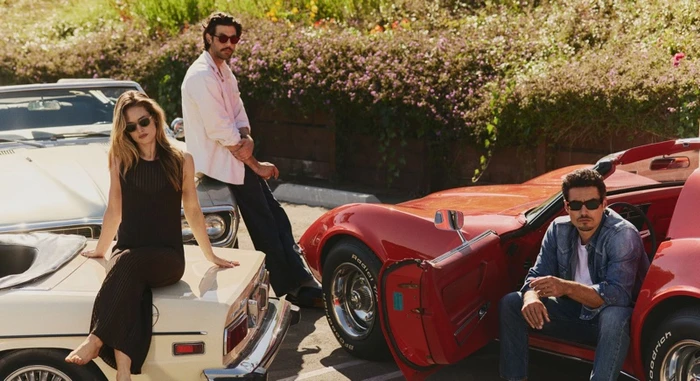 Three models wearing sunglasses posing with classic convertible cars in an outdoor parking lot setting.