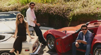 Three models wearing sunglasses posing with classic convertible cars in an outdoor parking lot setting.