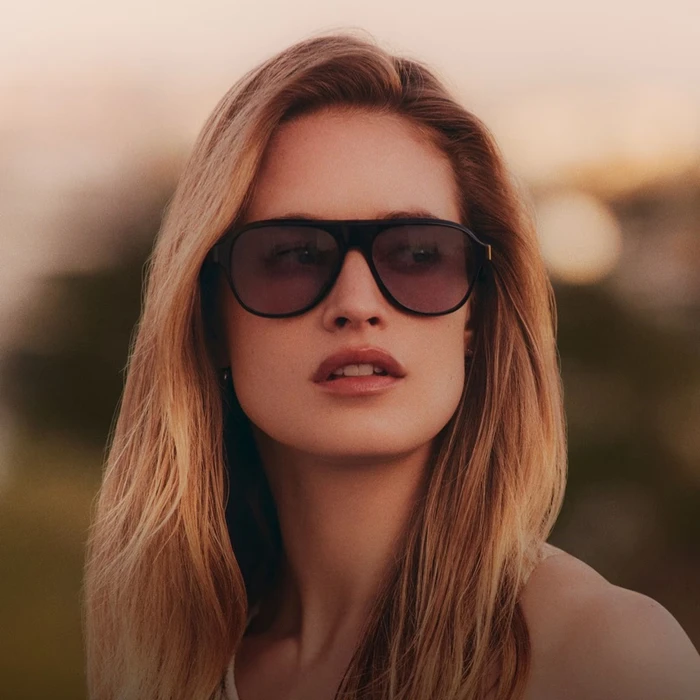 Model wearing dark sunglasses with light brown hair in warm sunset lighting for Oliver Peoples eyewear campaign.