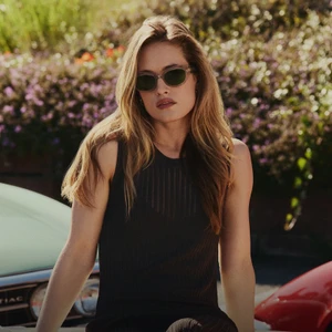Model wearing dark sunglasses and a dark sleeveless top outdoors near cars with blooming bushes behind her.
