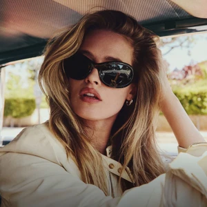 Model wearing dark sunglasses and a light jacket seated inside a vehicle with a blurred background