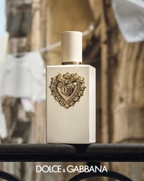 White perfume bottle with ornate gold heart emblem resting on a dark railing against a blurred street backdrop.