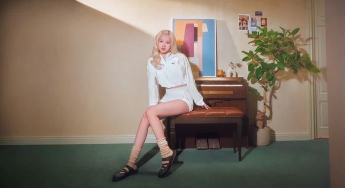ROSÉ seated on a leather piano bench in a retro living room wearing a white Puma tracksuit and black flats with socks
