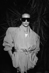 Rosie Huntington-Whiteley wearing oversized sunglasses and a belted jacket against dark foliage for FWRD Summer 2026.
