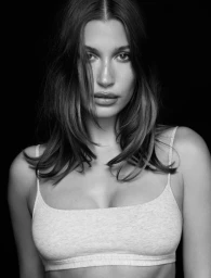 Hailey Bieber wearing a white cotton tank top against a dark background in a black and white portrait.