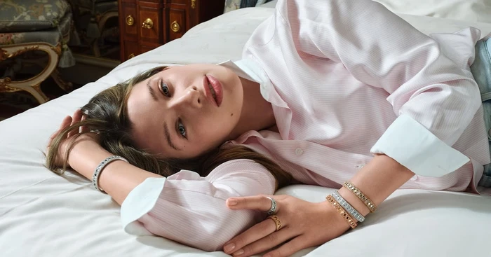 Ever Anderson reclining on white bedsheets wearing a striped pink shirt and Dior jewellery for My Dior